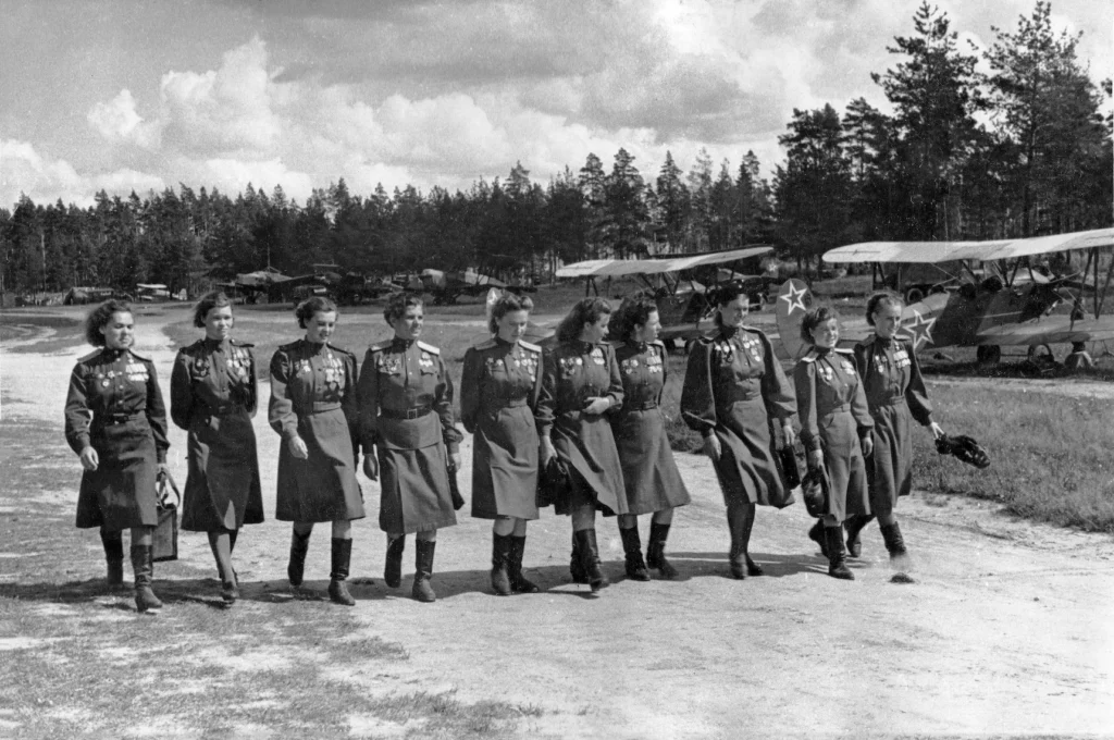 Grupo de aviadoras soviéticas das Bruxas da Noite caminhando em um campo de aviação com biplanos Polikarpov Po-2 ao fundo.